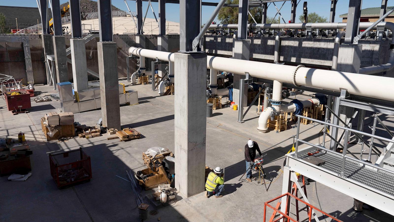 Two helmeted workers operate equipment amid concrete columns, large pipes, scaffolding, pallets, and construction materials at an industrial site.