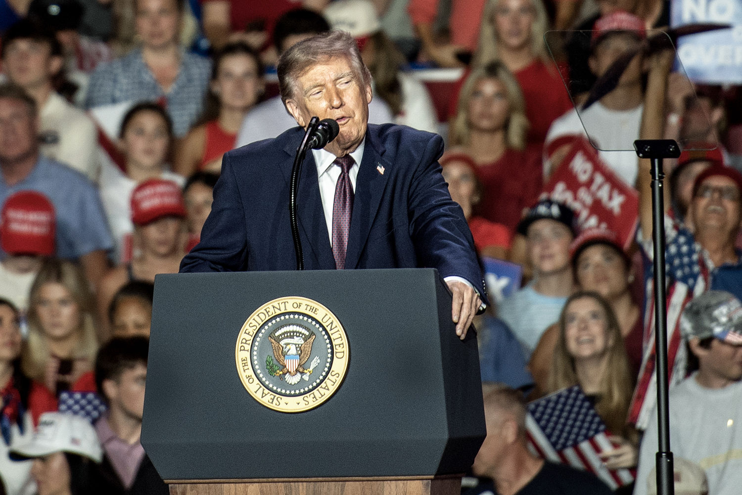 Trump speaks behind a presidential-seal lectern as supporters and a teleprompter fill the rally venue.
