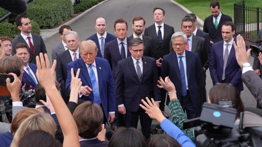 Trump stands among suited officials while reporters raise their hands and cameras outside.