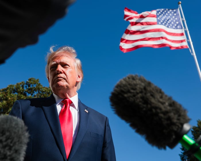 Trump stands outdoors in a dark suit and red tie, with microphones in the foreground and an American flag overhead.