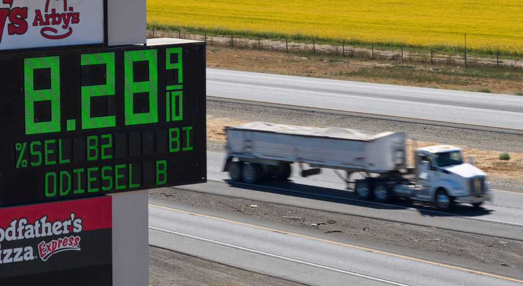 A truck passes a roadside fuel price sign displaying green digital numbers beside a highway.