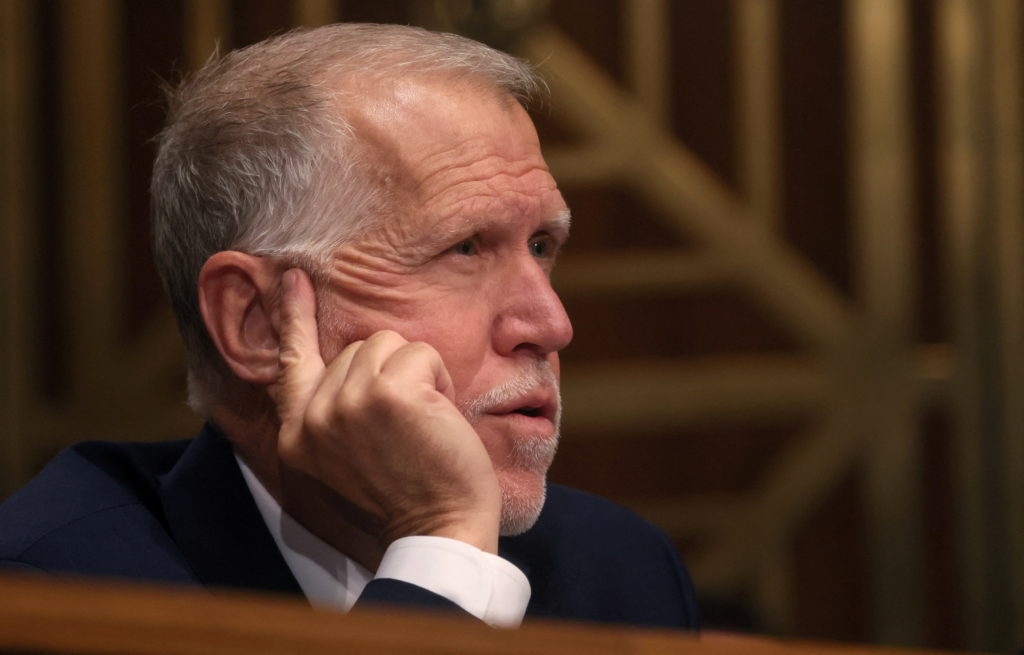 A gray-haired man in a dark suit sits with his hand against his cheek and a finger beside his ear.
