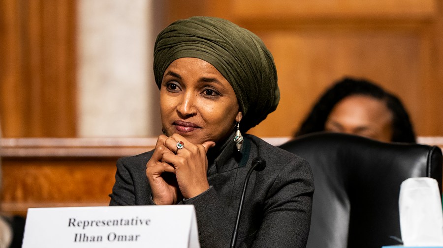 Ilhan Omar sits behind a nameplate and microphone, wearing a green headwrap with her hands clasped beneath her chin.