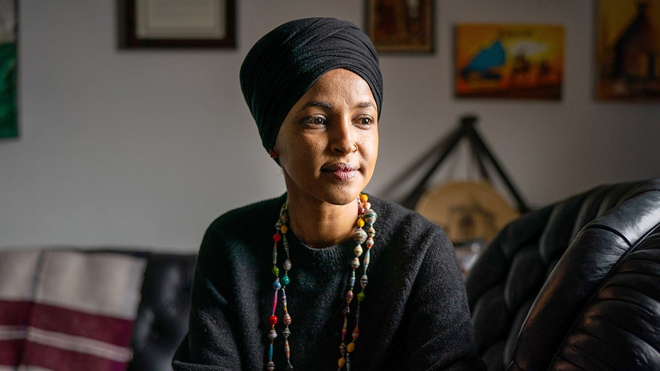 Omar sits on a black leather sofa wearing a black headwrap, dark sweater and multicolored beaded necklaces.