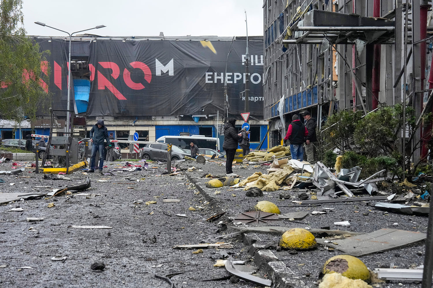 People stand on a debris-strewn street beside a damaged building, its panels, glass and insulation scattered across the wet pavement.