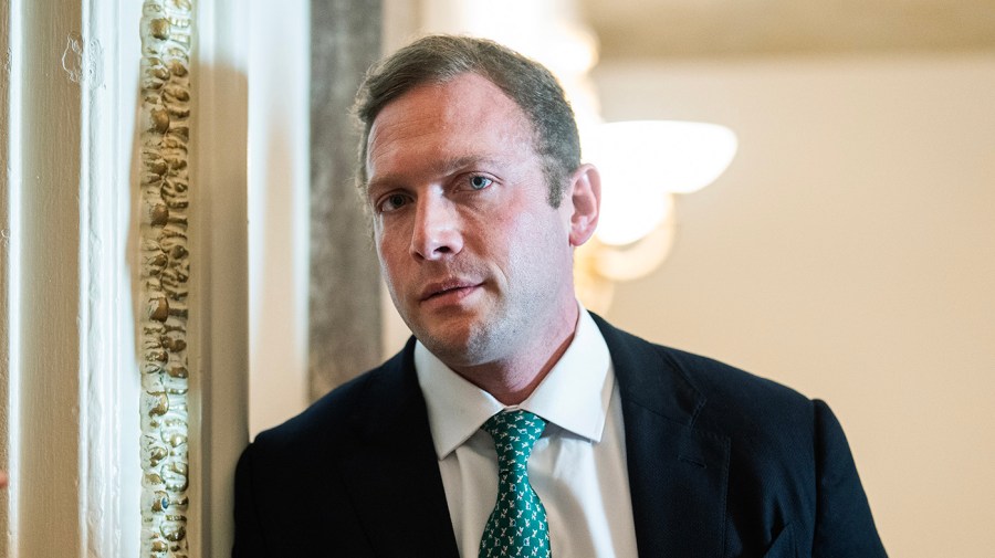 Rep. Max Miller, in a dark suit, white shirt and green patterned tie, stands beside an ornate gilded doorframe.