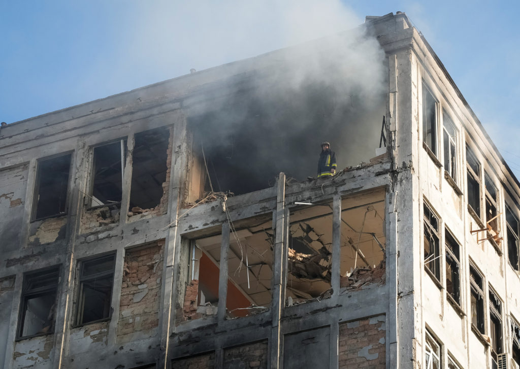 A firefighter stands on an exposed upper floor of a heavily damaged building as smoke pours from its shattered windows into a blue sky.