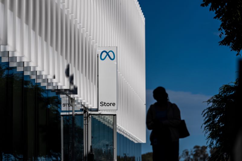 A pedestrian in silhouette walks past a Meta store, its white sign showing the blue Meta logo, under a deep blue sky.