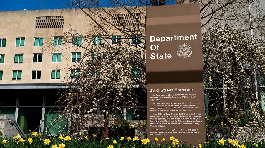 The Department of State sign at the 23rd Street entrance of its Washington headquarters, behind flowering trees and yellow daffodils.