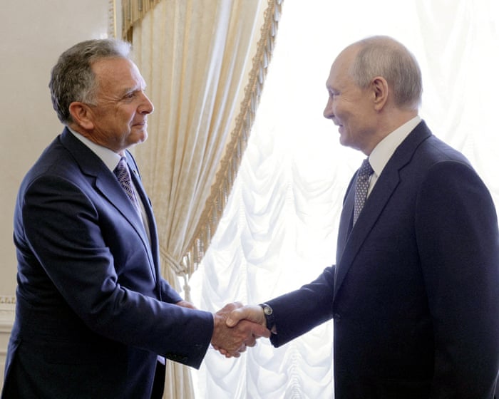 Steve Witkoff and Vladimir Putin, both in dark suits, shake hands in front of cream curtains.