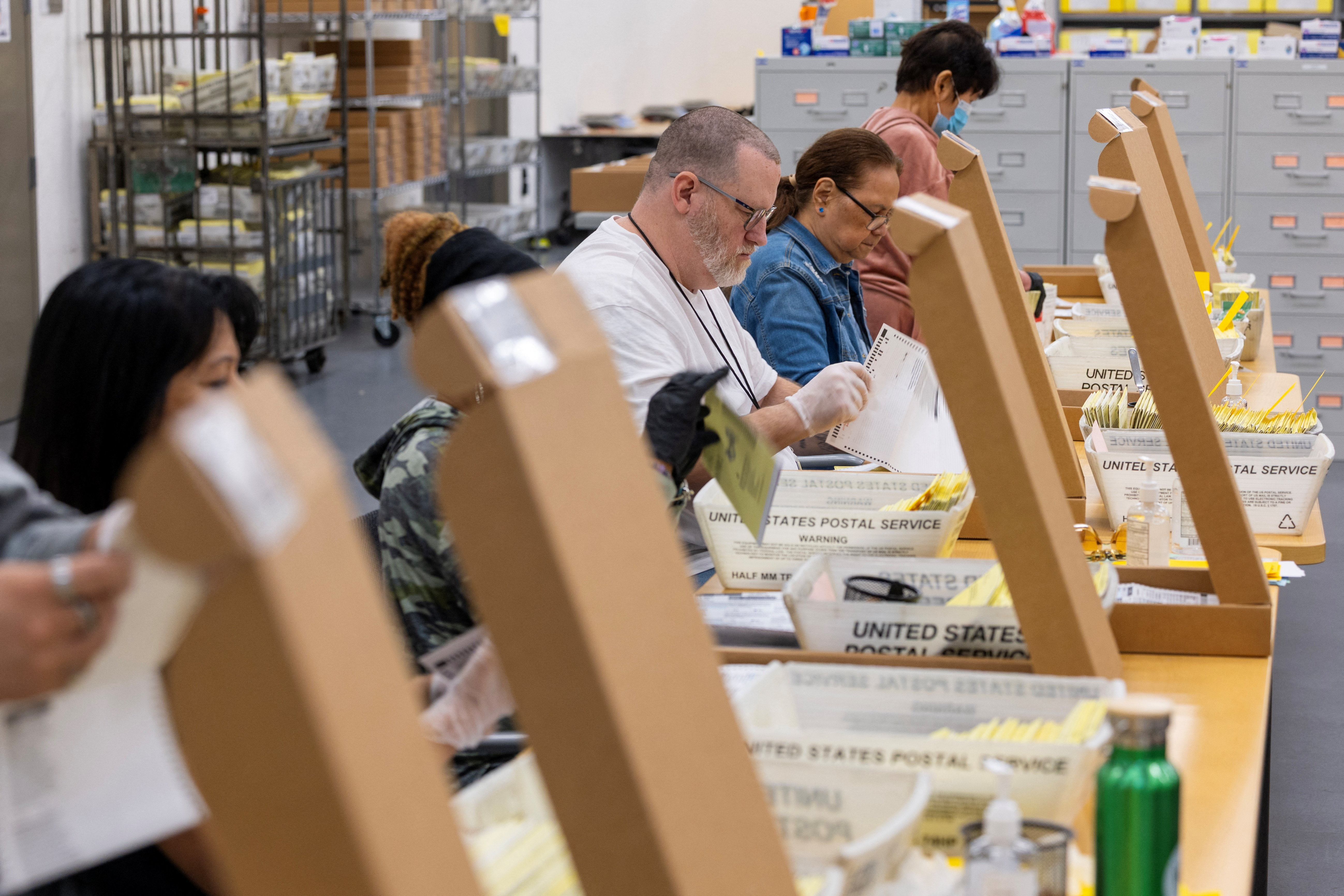 Election workers in gloves handle mail ballots at a long table lined with cardboard dividers and U.S. Postal Service trays.